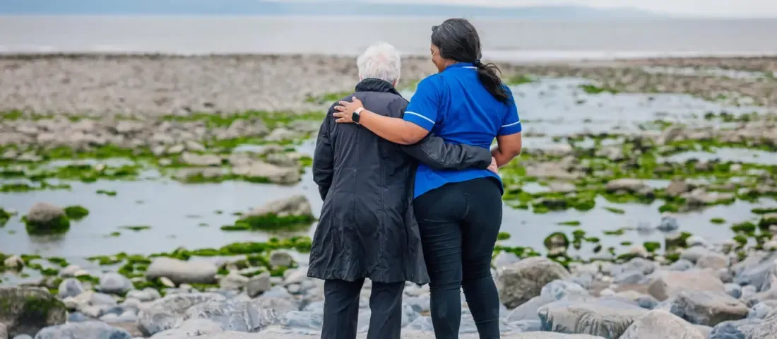 Caremark carer supporting an elderly person during a walk by the seaside